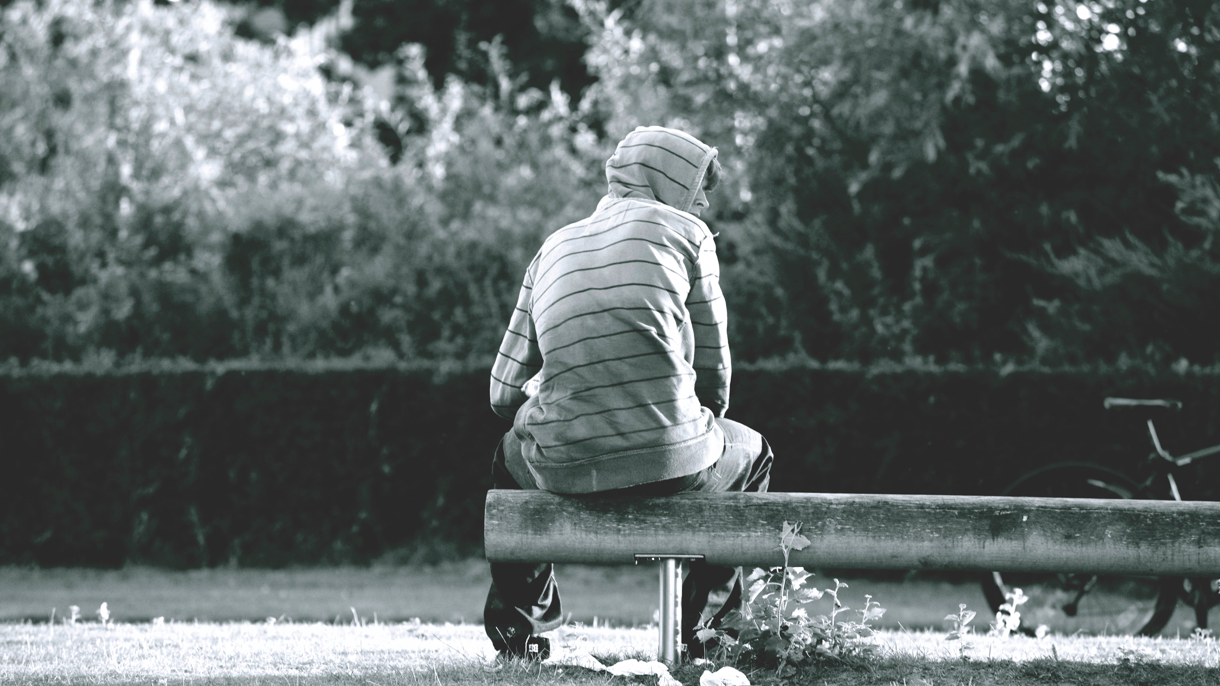 a boy is sitting alone on the bench  in the garden. he wear white coat and jeans. he looking at the his right side.