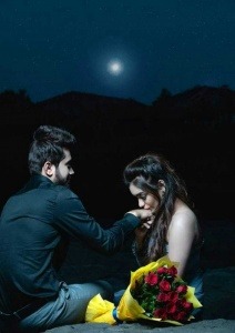 moon is going down and hills are blur in background. boy and girl are sitting on the big stone. boy wear black shirt and gray pent. girl is in gray colour dress. red roses are in yelow colour plastic. girl is kissing on boy's hand.